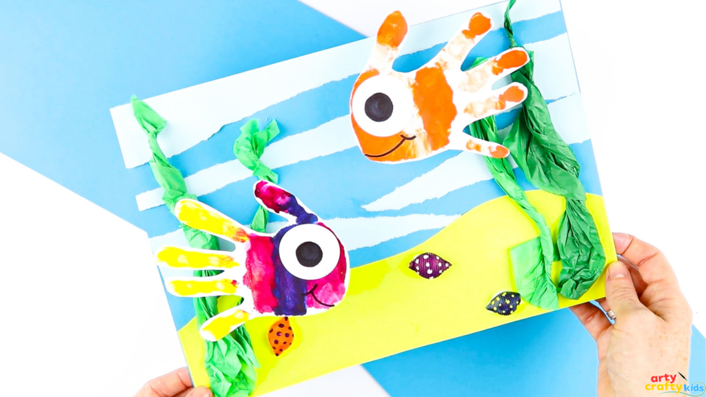 Fish handprint craft with torn paper water background, tissue paper seaweed, and painted pasta shells for texture.