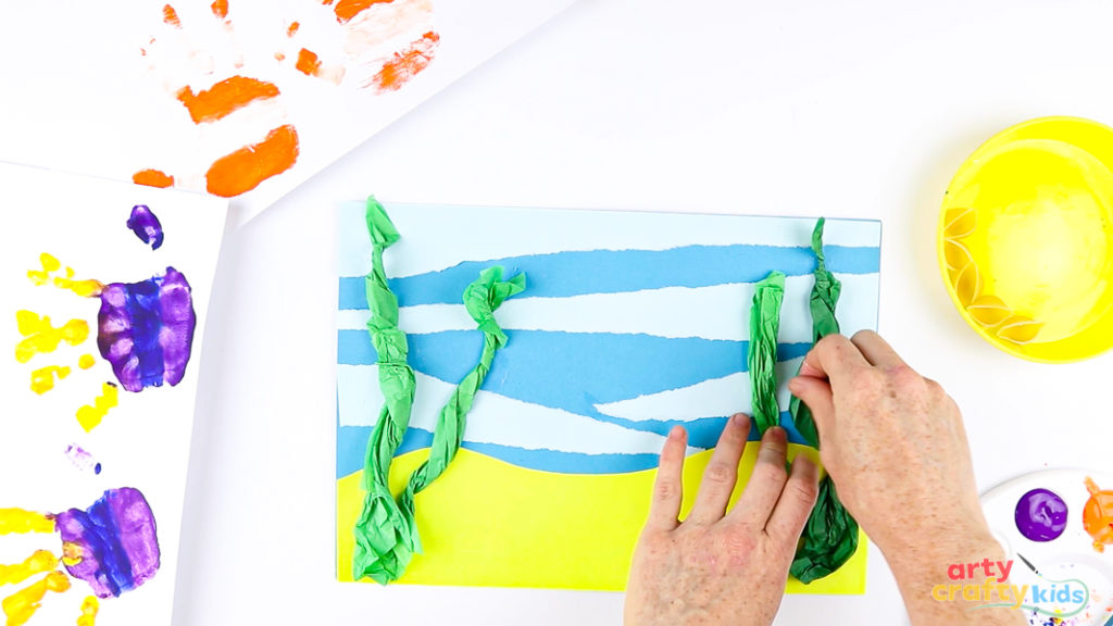 Image of a hand gluing green tissue paper to the ocean scene.