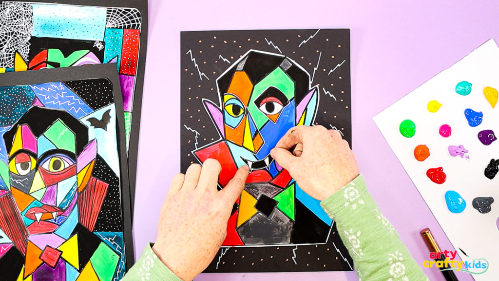 Assembling the Picasso Dracula collage with brightly painted face pieces arranged in a Cubist layout on black paper.